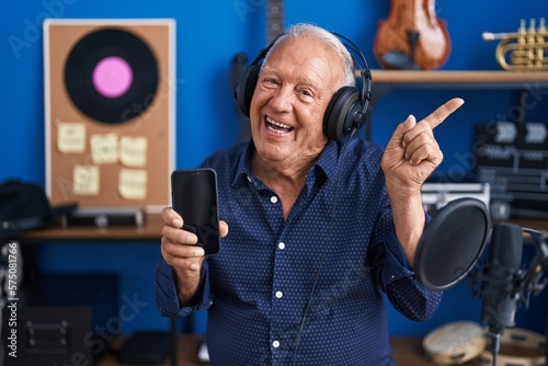 Senior man with grey hair showing smartphone screen at music studio smiling happy pointing with hand and finger to the side