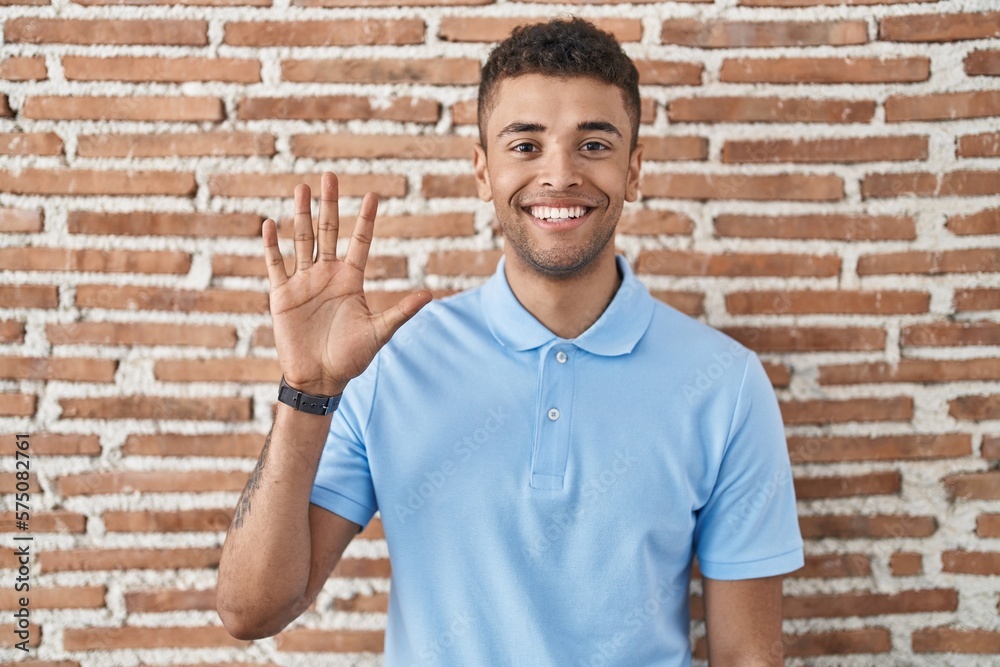 Brazilian young man standing over brick wall showing and pointing up with fingers number five while smiling confident and happy. Brazilian young man standing over brick wall showing and pointing up with fingers number five while smiling confident and happy.