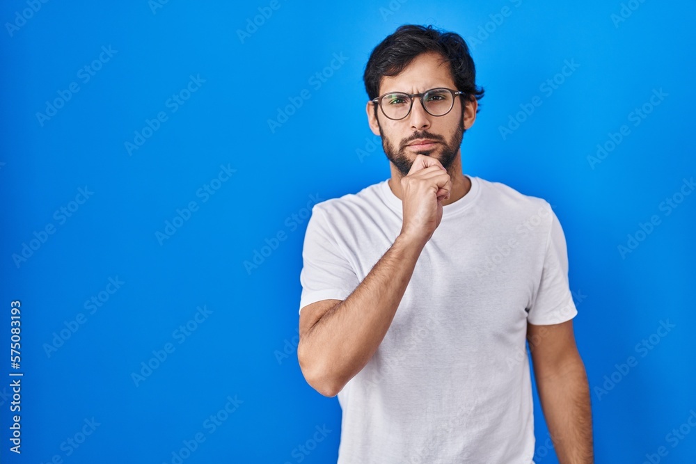 Handsome latin man standing over blue background looking confident at the camera with smile with crossed arms and hand raised on chin. thinking positive. Handsome latin man standing over blue background looking confident at the camera with smile with crossed arms and hand raised on chin. thinking positive.
