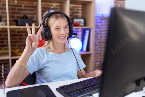 Young caucasian woman playing video games wearing headphones showing and pointing up with fingers number three while smiling confident and happy.