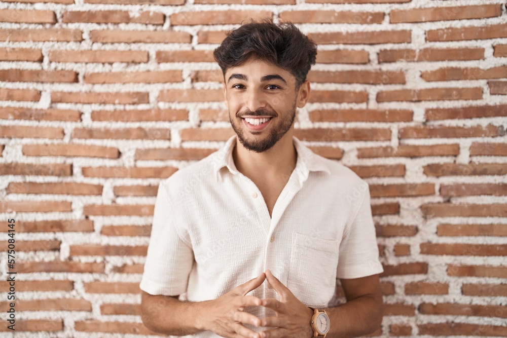 Arab man with beard standing over bricks wall background hands together and fingers crossed smiling relaxed and cheerful. success and optimistic
