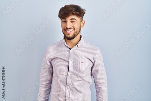 Arab man with beard standing over blue background winking looking at the camera with sexy expression, cheerful and happy face.