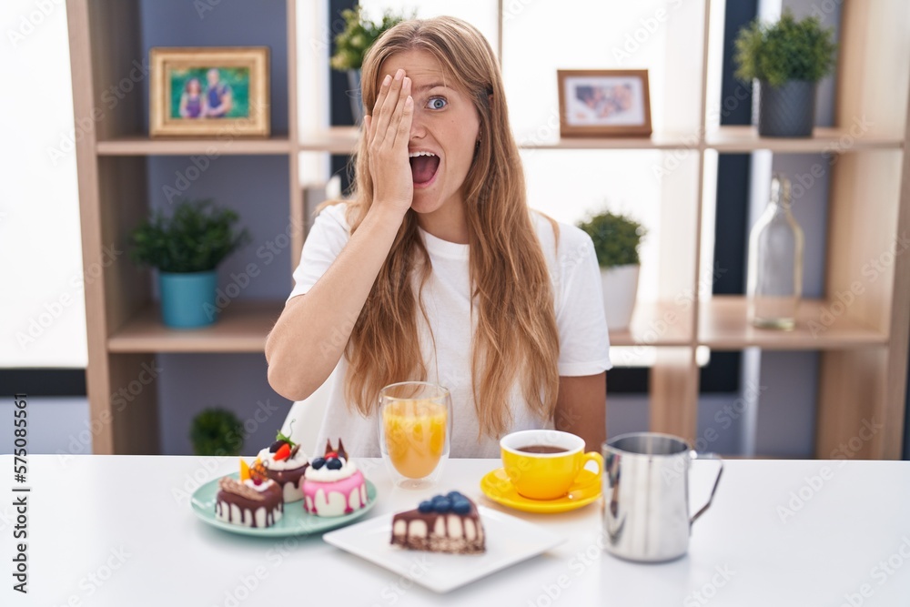 Young caucasian woman eating pastries t for breakfast covering one eye with hand, confident smile on face and surprise emotion.