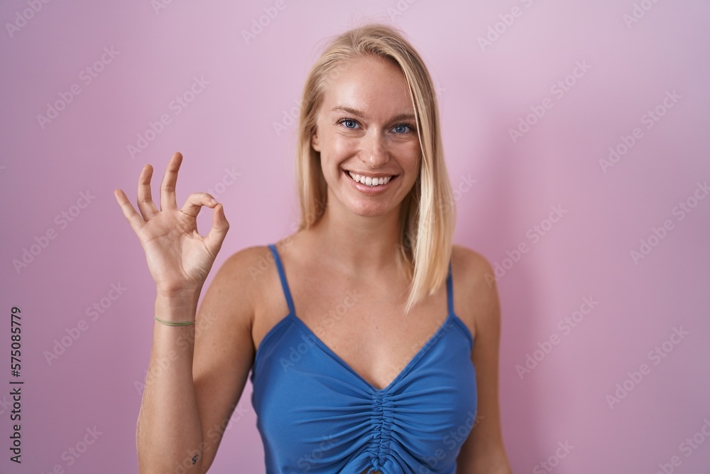 Young caucasian woman standing over pink background smiling positive doing ok sign with hand and fingers. successful expression. Young caucasian woman standing over pink background smiling positive doing ok sign with hand and fingers. successful expression.