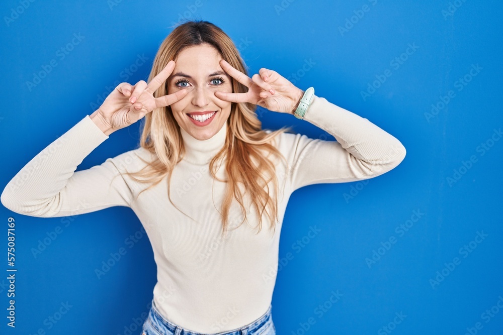 Young caucasian woman standing over blue background doing peace symbol with fingers over face, smiling cheerful showing victory Young caucasian woman standing over blue background doing peace symbol with fingers over face, smiling cheerful showing victory