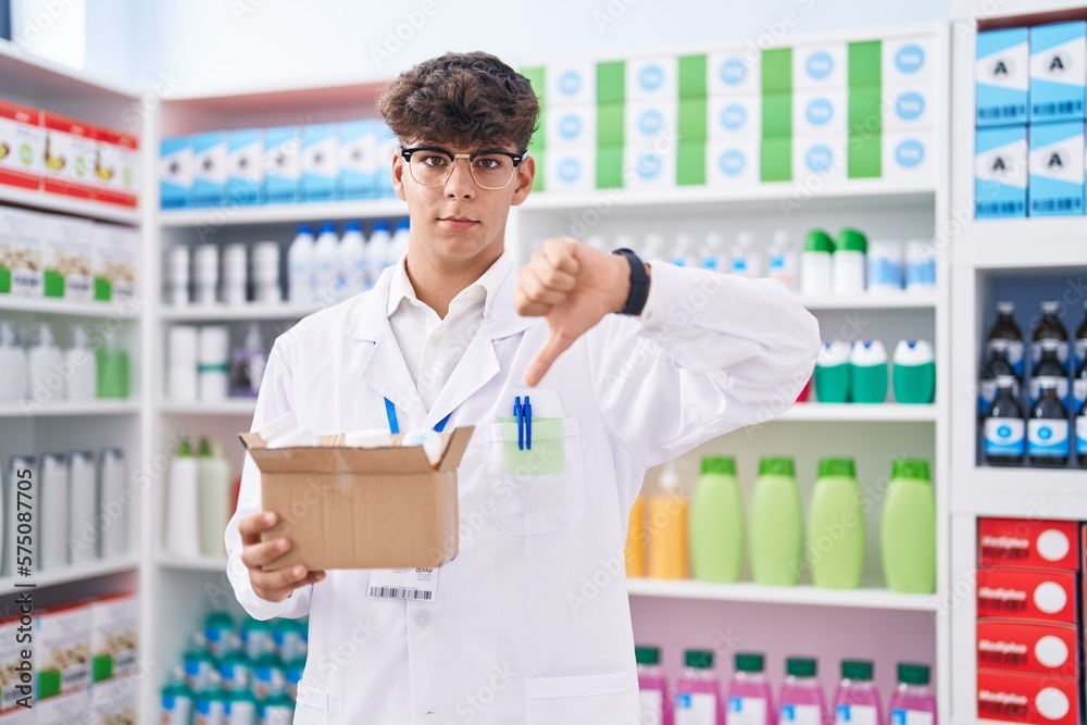 Hispanic teenager working at pharmacy drugstore holding box with pills with angry face, negative sign showing dislike with thumbs down, rejection concept Hispanic teenager working at pharmacy drugstore holding box with pills with angry face, negative sign showing dislike with thumbs down, rejection concept