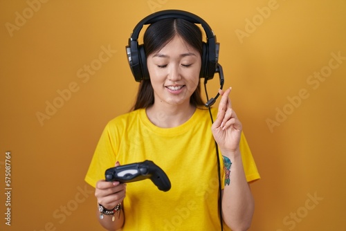 Chinese young woman playing video game holding controller gesturing finger crossed smiling with hope and eyes closed. luck and superstitious concept.