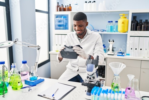 Young african american man wearing scientist uniform holding virtual reality glasses at laboratory