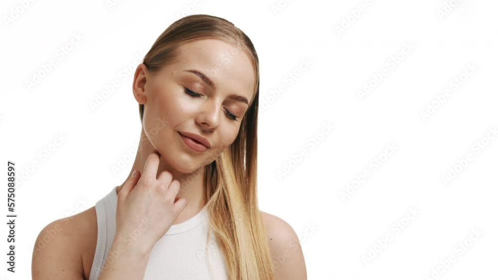 Good-looking fit European young woman with long blond hair in ponytail in white top turns to the camera touching her face and smiling wide for it on white background Good-looking fit European young woman with long blond hair in ponytail in white top turns to the camera touching her face and smiling wide for it on white background