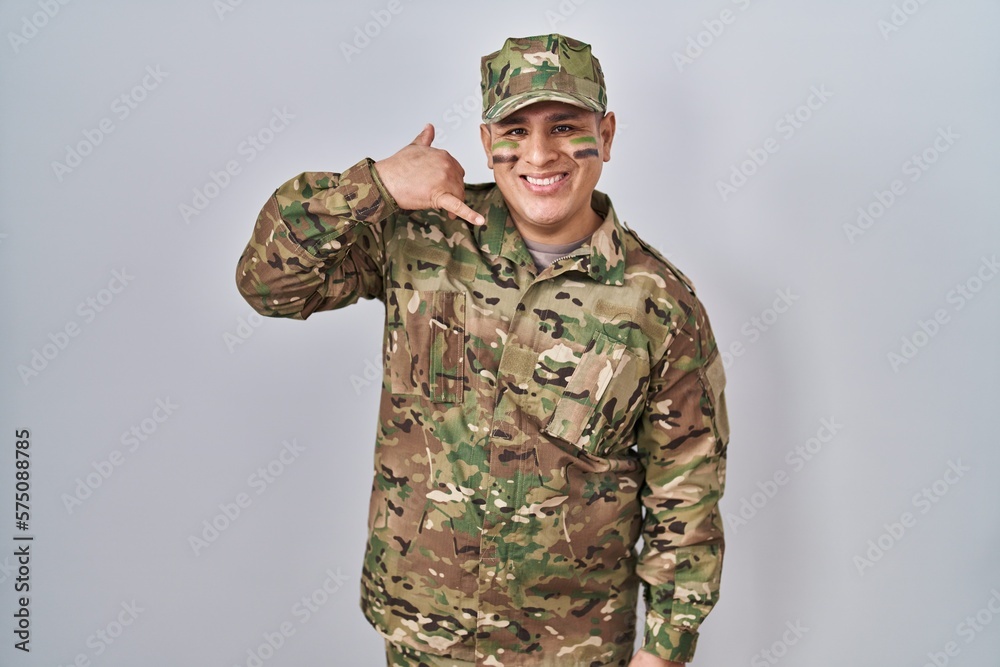 Hispanic young man wearing camouflage army uniform smiling doing phone gesture with hand and fingers like talking on the telephone. communicating concepts. Hispanic young man wearing camouflage army uniform smiling doing phone gesture with hand and fingers like talking on the telephone. communicating concepts.
