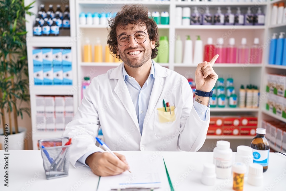Hispanic young man working at pharmacy drugstore with a big smile on face, pointing with hand finger to the side looking at the camera.
