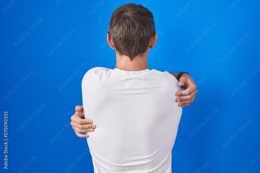 Caucasian blond man standing over blue background hugging oneself happy and positive from backwards. self love and self care Caucasian blond man standing over blue background hugging oneself happy and positive from backwards. self love and self care