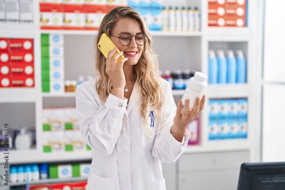Young woman pharmacist talking on smartphone holding pills bottle at pharmacy