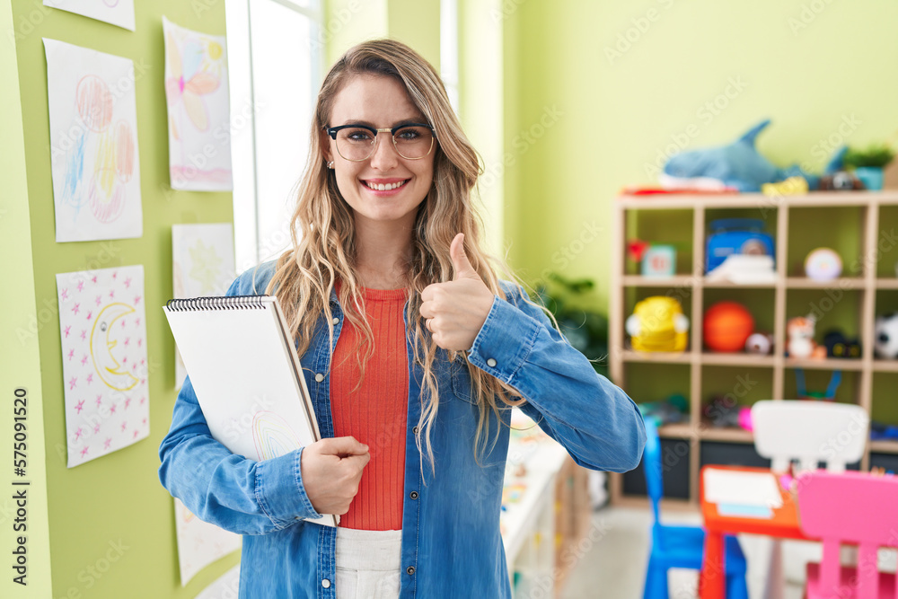 Young caucasian woman working as teacher at kindergarten smiling happy and positive, thumb up doing excellent and approval sign - obrazy, fototapety, plakaty 