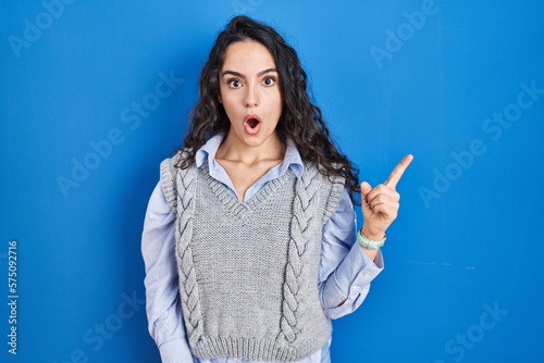 Young brunette woman standing over blue background surprised pointing with finger to the side, open mouth amazed expression.