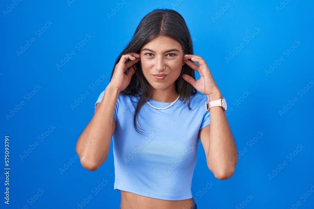 Brunette young woman standing over blue background covering ears with fingers with annoyed expression for the noise of loud music. deaf concept. Brunette young woman standing over blue background covering ears with fingers with annoyed expression for the noise of loud music. deaf concept.
