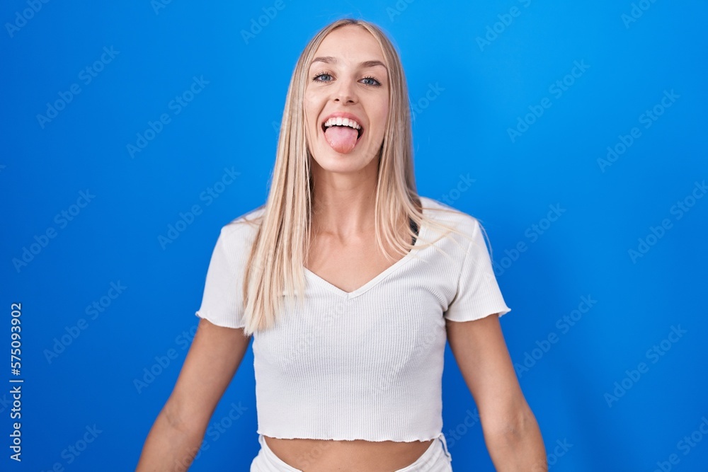 Young caucasian woman standing over blue background sticking tongue out happy with funny expression. emotion concept.