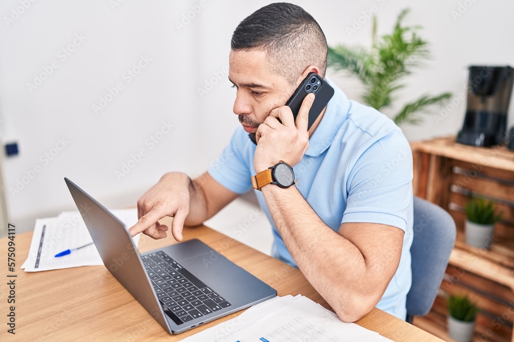 Young latin man business worker using laptop talking on smartphone at office Young latin man business worker using laptop talking on smartphone at office