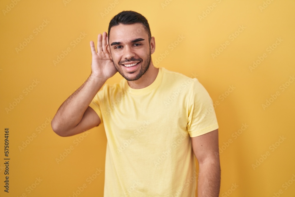 Young hispanic man standing over yellow background smiling with hand over ear listening an hearing to rumor or gossip. deafness concept. Young hispanic man standing over yellow background smiling with hand over ear listening an hearing to rumor or gossip. deafness concept.