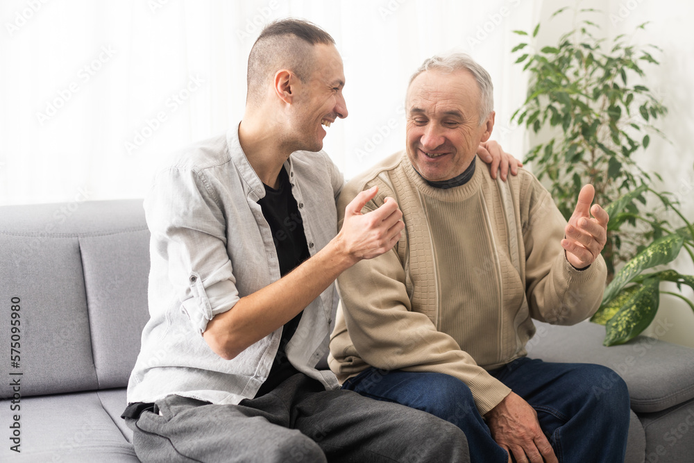 Happy two generations male family old senior mature father and smiling young adult grown son enjoying talking chatting bonding relaxing having friendly positive conversation sit on sofa at home. Happy two generations male family old senior mature father and smiling young adult grown son enjoying talking chatting bonding relaxing having friendly positive conversation sit on sofa at home.