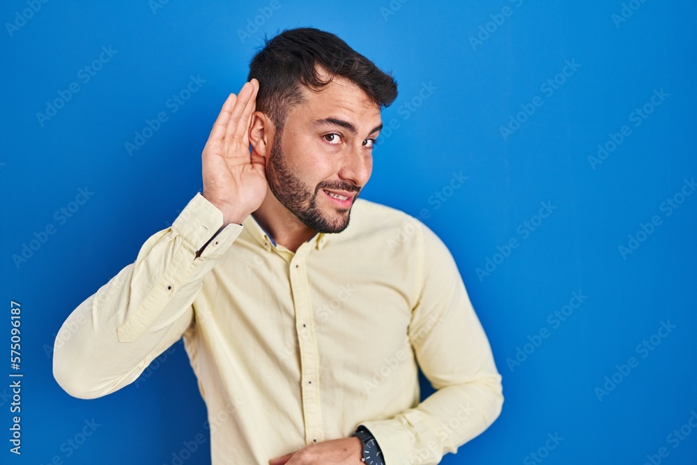Handsome hispanic man standing over blue background smiling with hand over ear listening an hearing to rumor or gossip. deafness concept. Handsome hispanic man standing over blue background smiling with hand over ear listening an hearing to rumor or gossip. deafness concept.