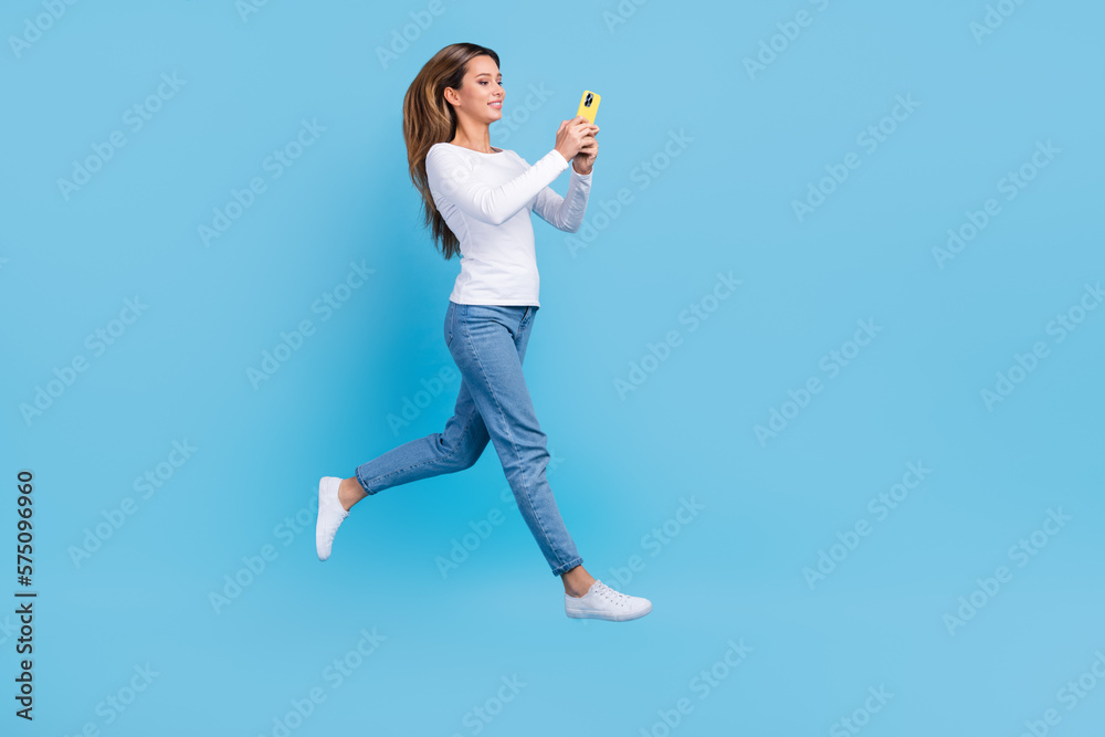 Fototapeta premium Full length photo of pretty shiny woman wear white shirt jumping high chatting device empty space isolated blue color background