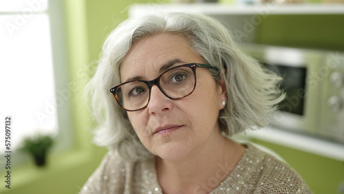 Middle age woman with grey hair sitting on table with relaxed expression at home