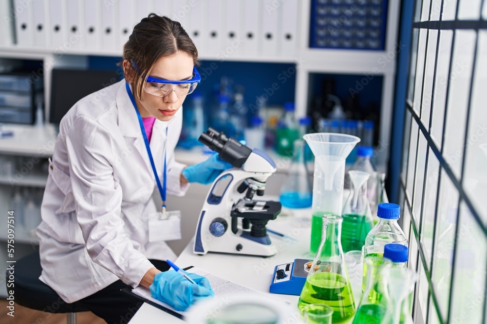 Fototapeta premium Young beautiful hispanic woman scientist using microscope writing on document at laboratory