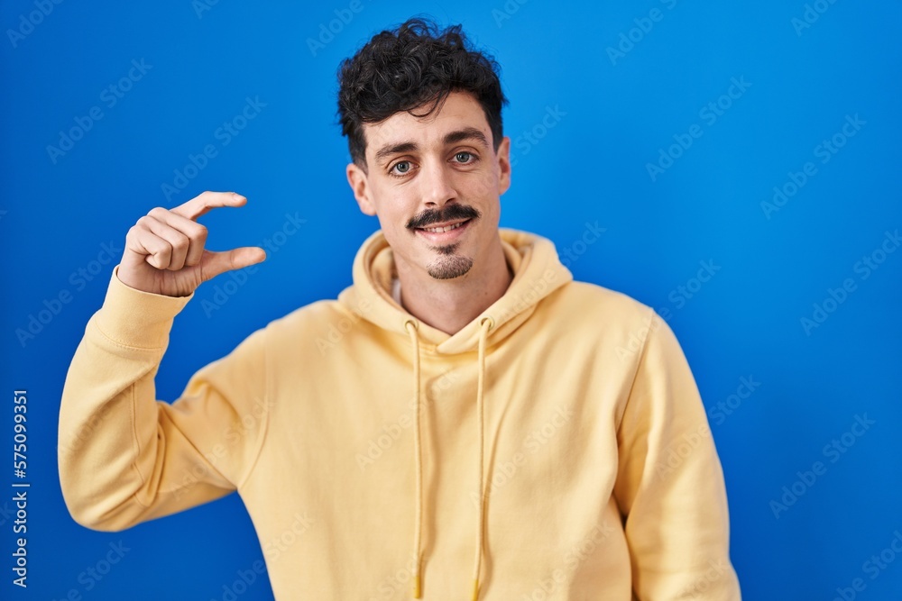 Hispanic man standing over blue background smiling and confident gesturing with hand doing small size sign with fingers looking and the camera. measure concept. Hispanic man standing over blue background smiling and confident gesturing with hand doing small size sign with fingers looking and the camera. measure concept.