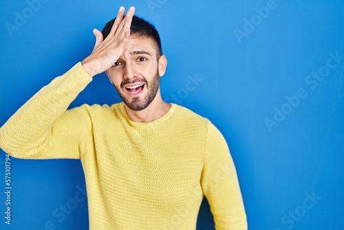 Hispanic man standing over blue background surprised with hand on head for mistake, remember error. forgot, bad memory concept.
