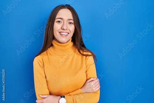 Young brunette woman standing over blue background happy face smiling with crossed arms looking at the camera. positive person.