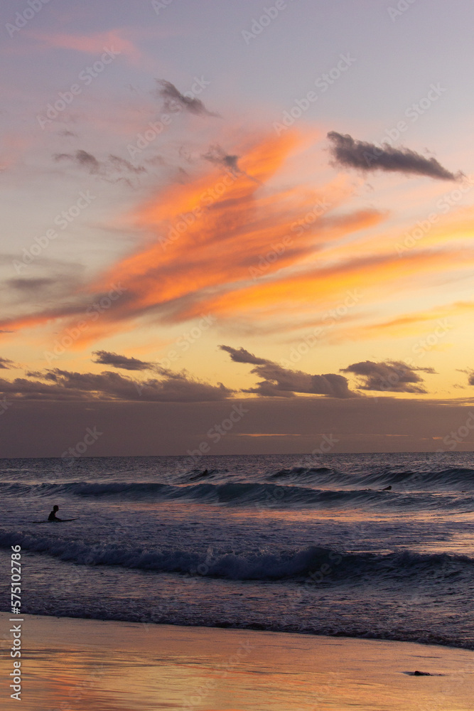 A beautiful sunset view of the beach with surfers in the water and orange clouds above in Fuerteventura
