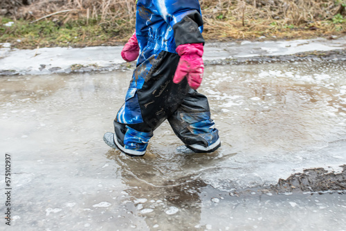 A small child walks through a dirty puddle in rubber boots in the spring with pieces of ice
