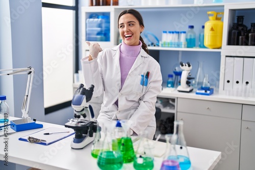 Young brunette woman working at scientist laboratory smiling with happy face looking and pointing to the side with thumb up.