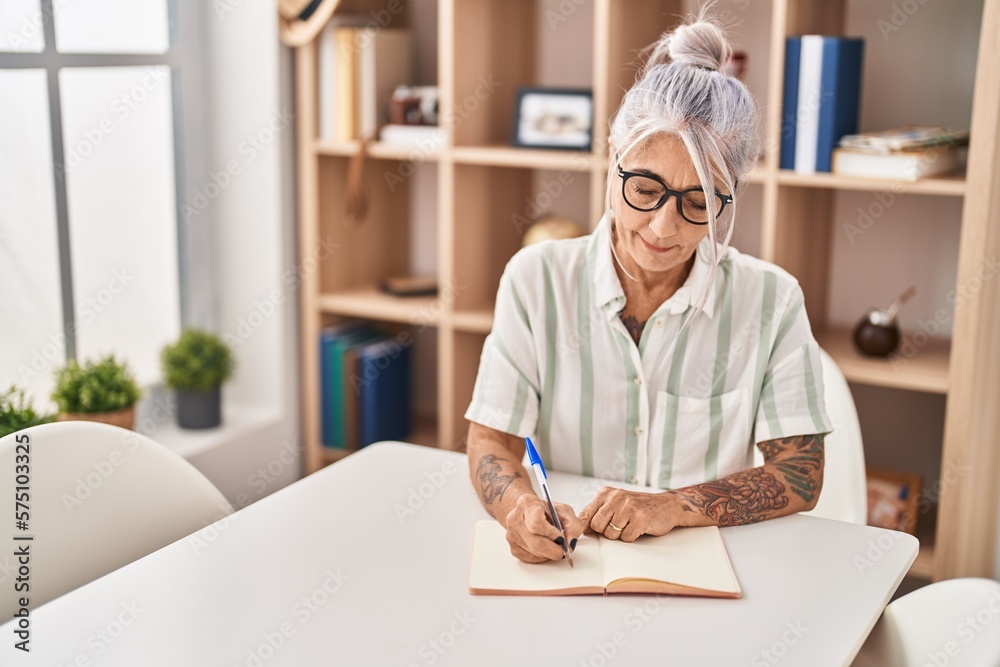 Middle age grey-haired woman writing on notebook sitting on table at home Middle age grey-haired woman writing on notebook sitting on table at home
