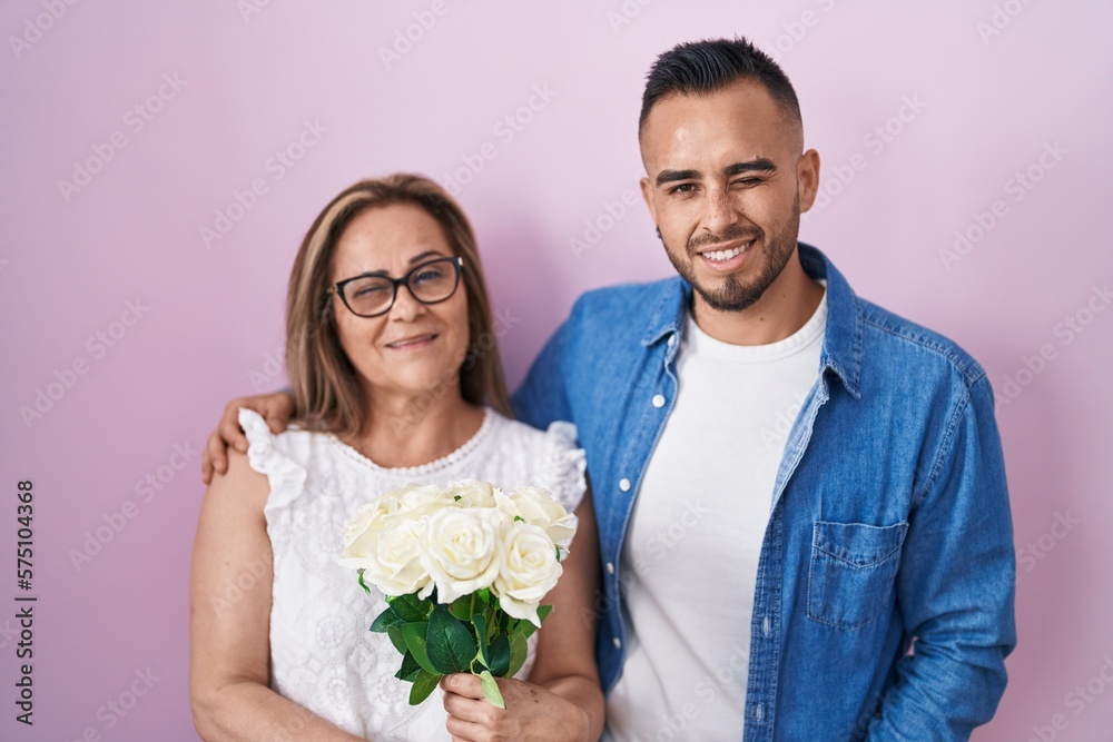Hispanic mother and son together holding bouquet of white flowers winking looking at the camera with sexy expression, cheerful and happy face. Hispanic mother and son together holding bouquet of white flowers winking looking at the camera with sexy expression, cheerful and happy face.