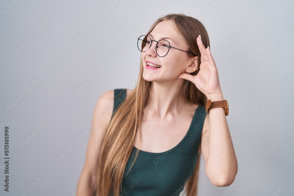 Young caucasian woman standing over white background smiling with hand over ear listening an hearing to rumor or gossip. deafness concept.