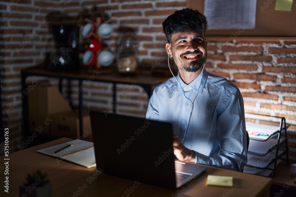 Young hispanic man with beard working at the office at night looking away to side with smile on face, natural expression. laughing confident. Young hispanic man with beard working at the office at night looking away to side with smile on face, natural expression. laughing confident.