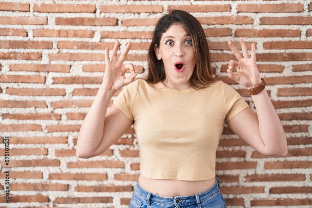 Young brunette woman standing over bricks wall looking surprised and shocked doing ok approval symbol with fingers. crazy expression Young brunette woman standing over bricks wall looking surprised and shocked doing ok approval symbol with fingers. crazy expression