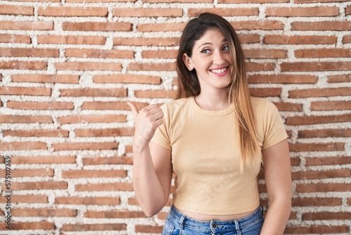 Young brunette woman standing over bricks wall smiling with happy face looking and pointing to the side with thumb up. © Krakenimages.com