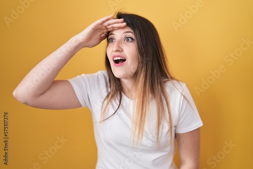Young brunette woman standing over yellow background very happy and smiling looking far away with hand over head. searching concept.