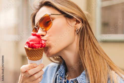 Close up happy young woman with delicious pink ice cream in waffle cone outdoors. Girl wear orange sunglasses in summer, spring or fall sunny day. Blonde female lick ice cream and laughing.