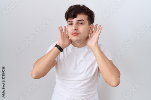 Young non binary man wearing casual white t shirt trying to hear both hands on ear gesture, curious for gossip. hearing problem, deaf © Krakenimages.com