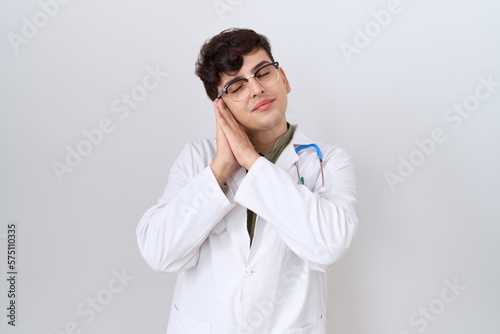 Young non binary man wearing doctor uniform and stethoscope sleeping tired dreaming and posing with hands together while smiling with closed eyes.