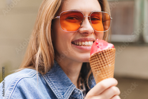 Close up happy young woman with delicious pink ice cream in waffle cone outdoors. Girl wear orange sunglasses in summer, spring or fall sunny day. Blonde female laughing and look at side.