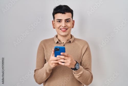 Non binary person using smartphone typing message smiling with a happy and cool smile on face. showing teeth.
