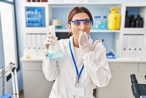 Young brunette woman working at scientist laboratory covering mouth with hand, shocked and afraid for mistake. surprised expression
