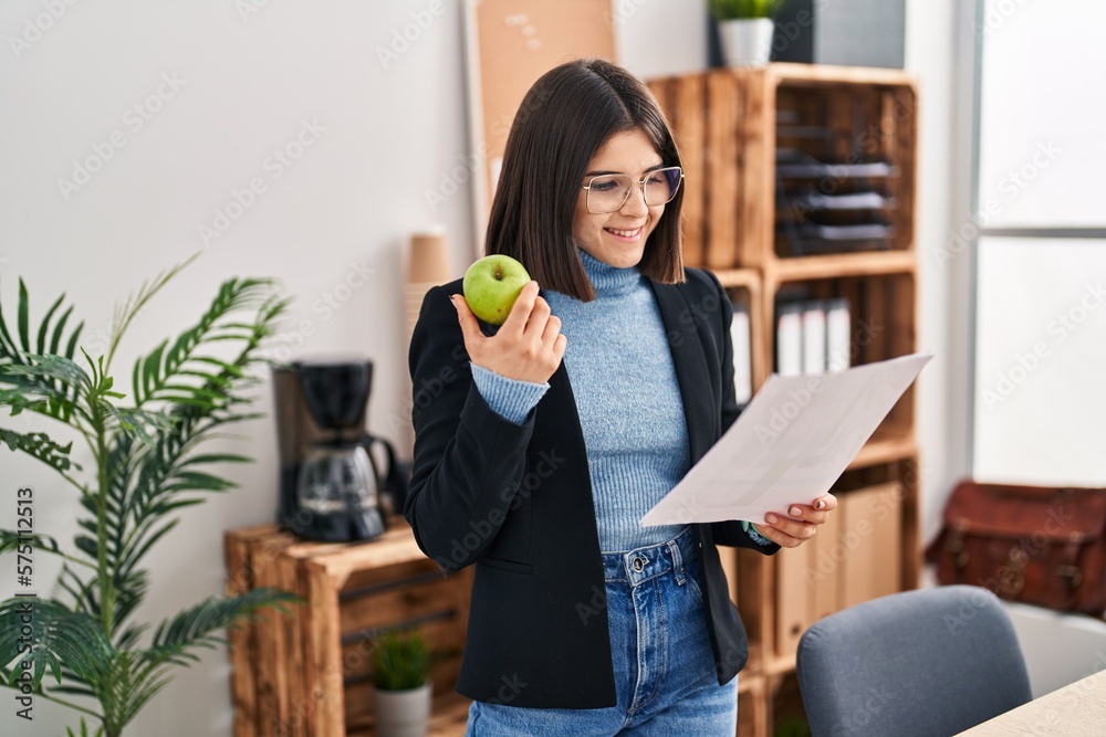 Young beautiful hispanic woman business worker reading document eating apple at office