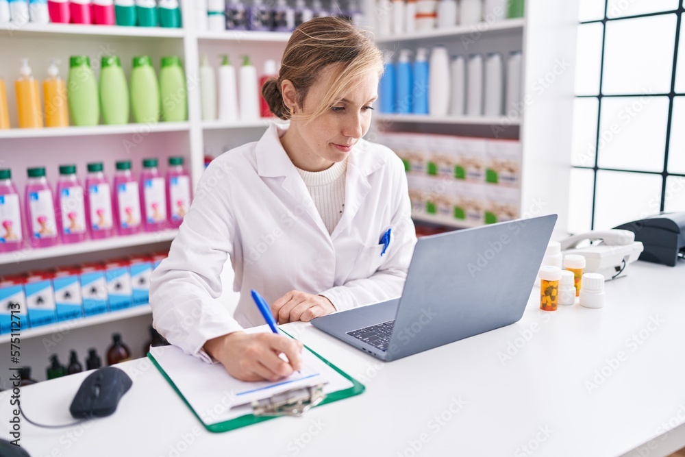 Young blonde woman pharmacist using laptop writing on document at pharmacy