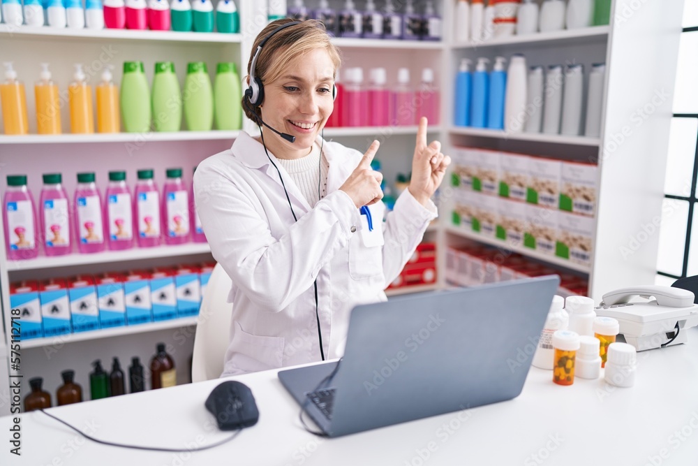 Young caucasian woman working at pharmacy drugstore using laptop smiling and looking at the camera pointing with two hands and fingers to the side.